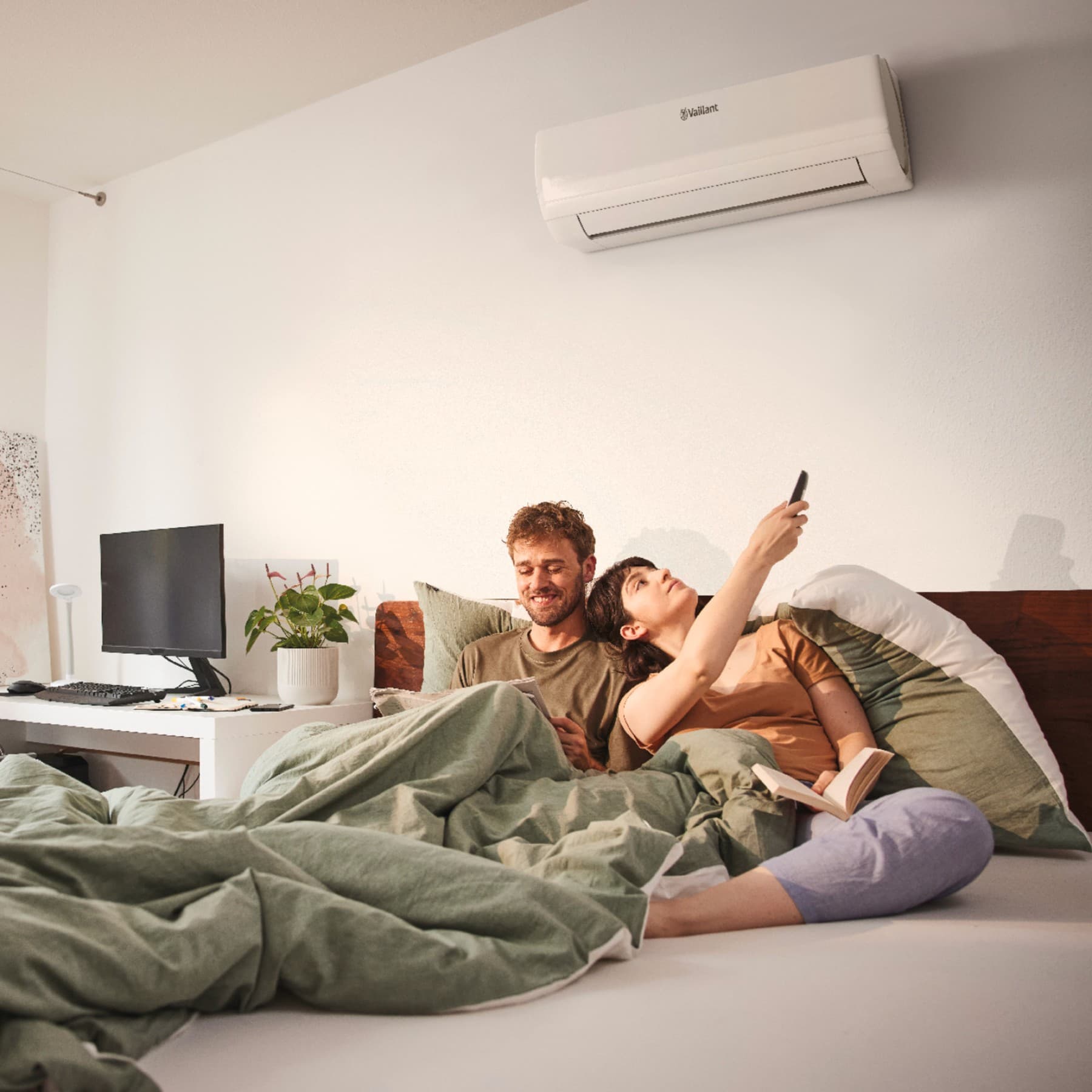 Le couple est encore au lit, sous des couvertures vertes ; la femme allume la climatisation à l'aide d'une télécommande. À l'arrière-plan, on aperçoit un bureau sur lequel se trouvent une plante, un écran et un clavier.