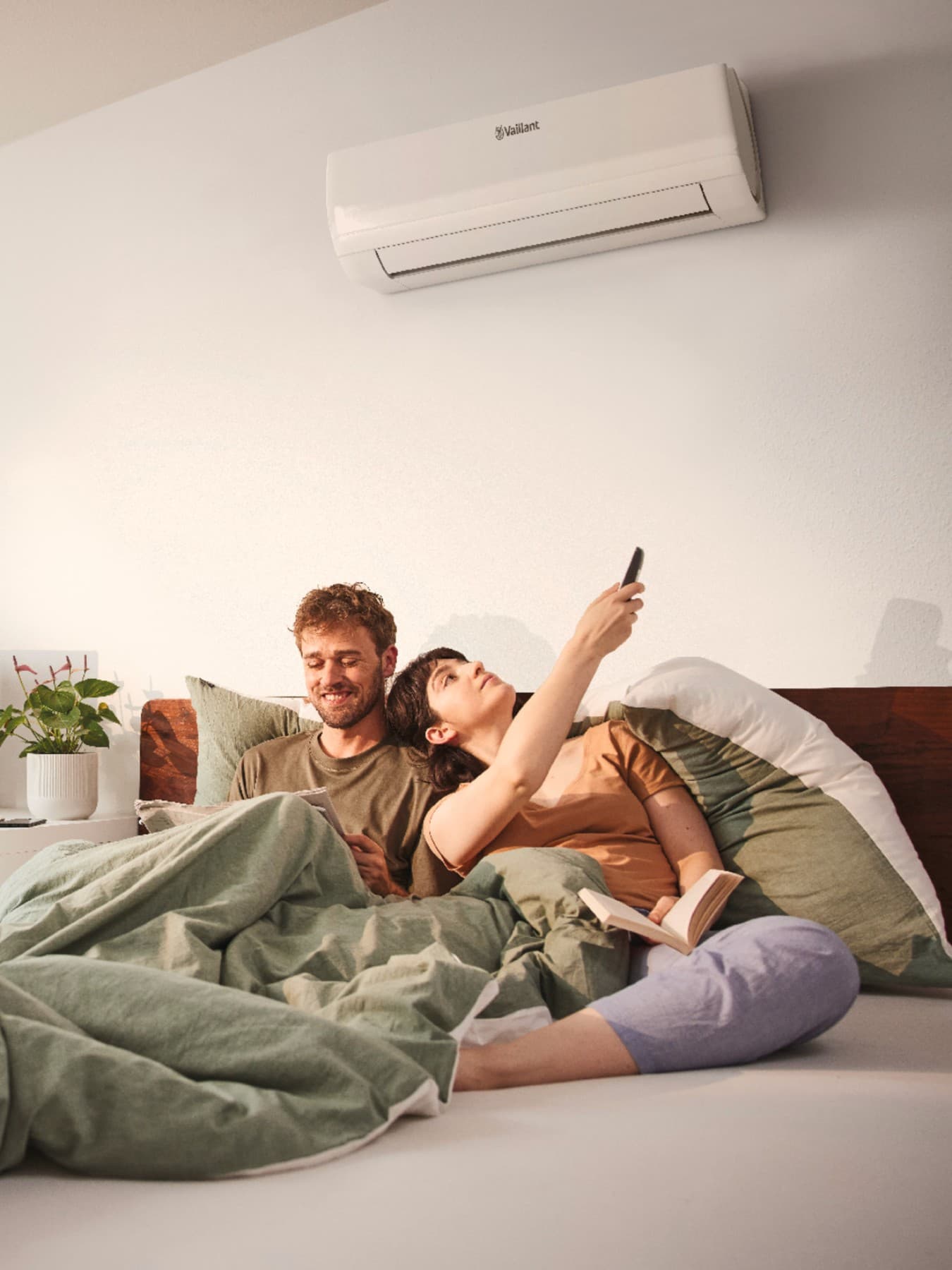 Le couple est encore au lit, sous des couvertures vertes ; la femme allume la climatisation à l'aide d'une télécommande. À l'arrière-plan, on aperçoit un bureau sur lequel se trouvent une plante, un écran et un clavier.