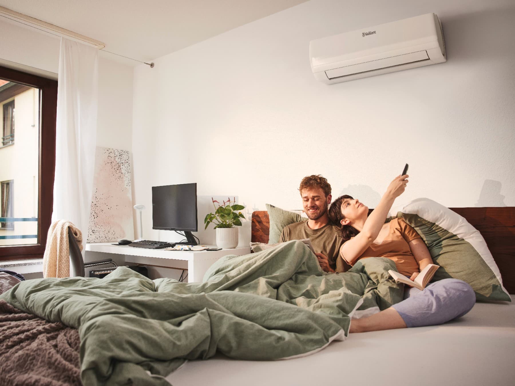 Le couple est au lit ; la femme pointe la télécommande vers le climatiseur pour l'allumer.