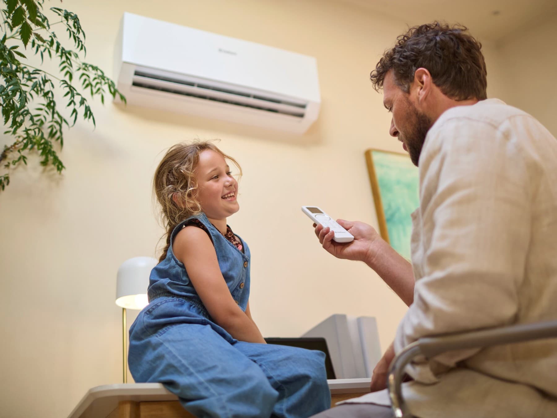 Une petite fille est assise sur un canapé, son père tient la télécommande de la climatisation