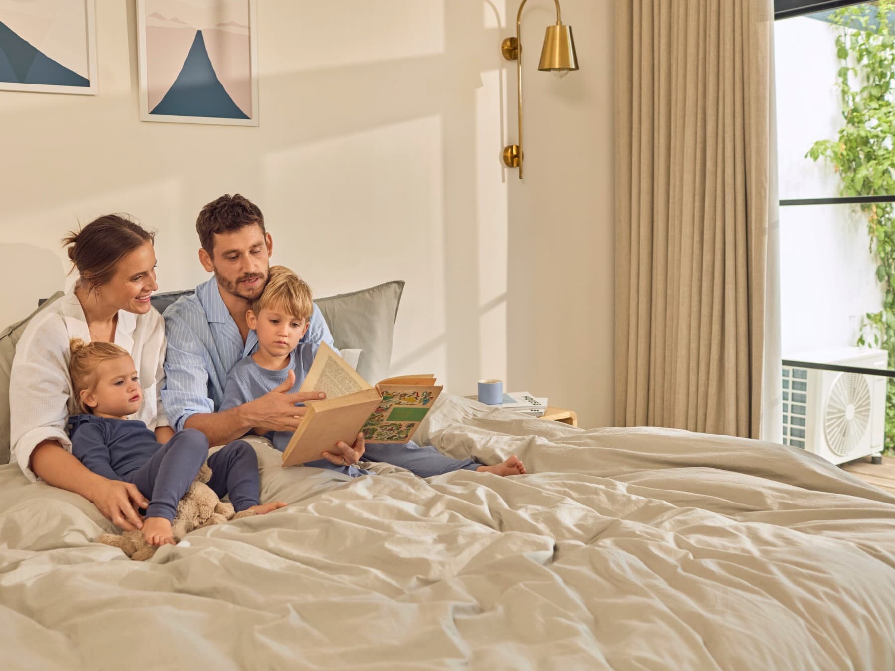 Koppel zit in bed. Er zitten twee kindjes op schoot waaraan ze voorlezen. Rechts zie je een raam met gordijnen waardoor je de buitenunit van een airco ziet