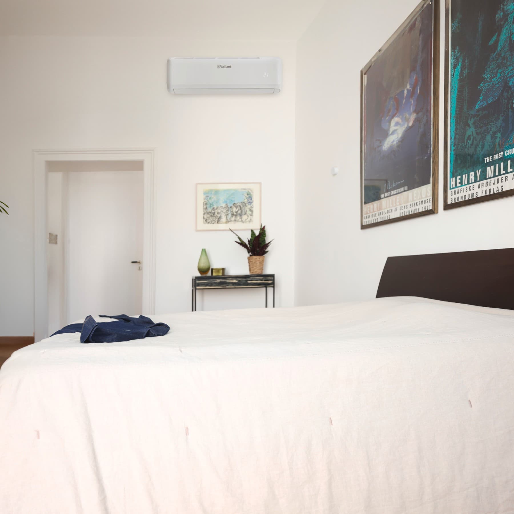 Chambre dans laquelle se trouve un t-shirt bleu posé sur un lit ; il y a des cadres accrochés au mur et une petite table sur laquelle sont posés une plante et un vase
