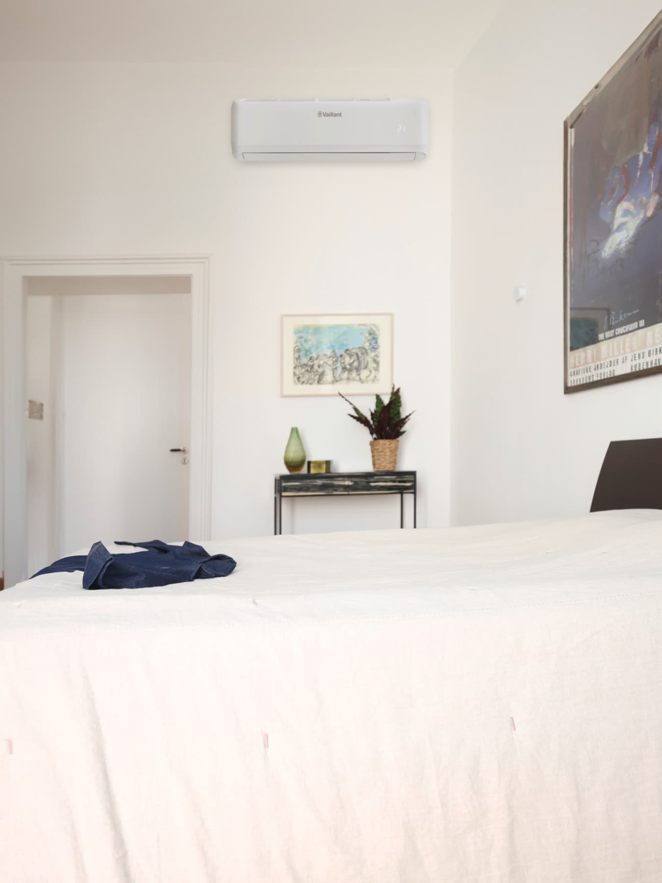 Chambre dans laquelle se trouve un t-shirt bleu posé sur un lit ; il y a des cadres accrochés au mur et une petite table sur laquelle sont posés une plante et un vase