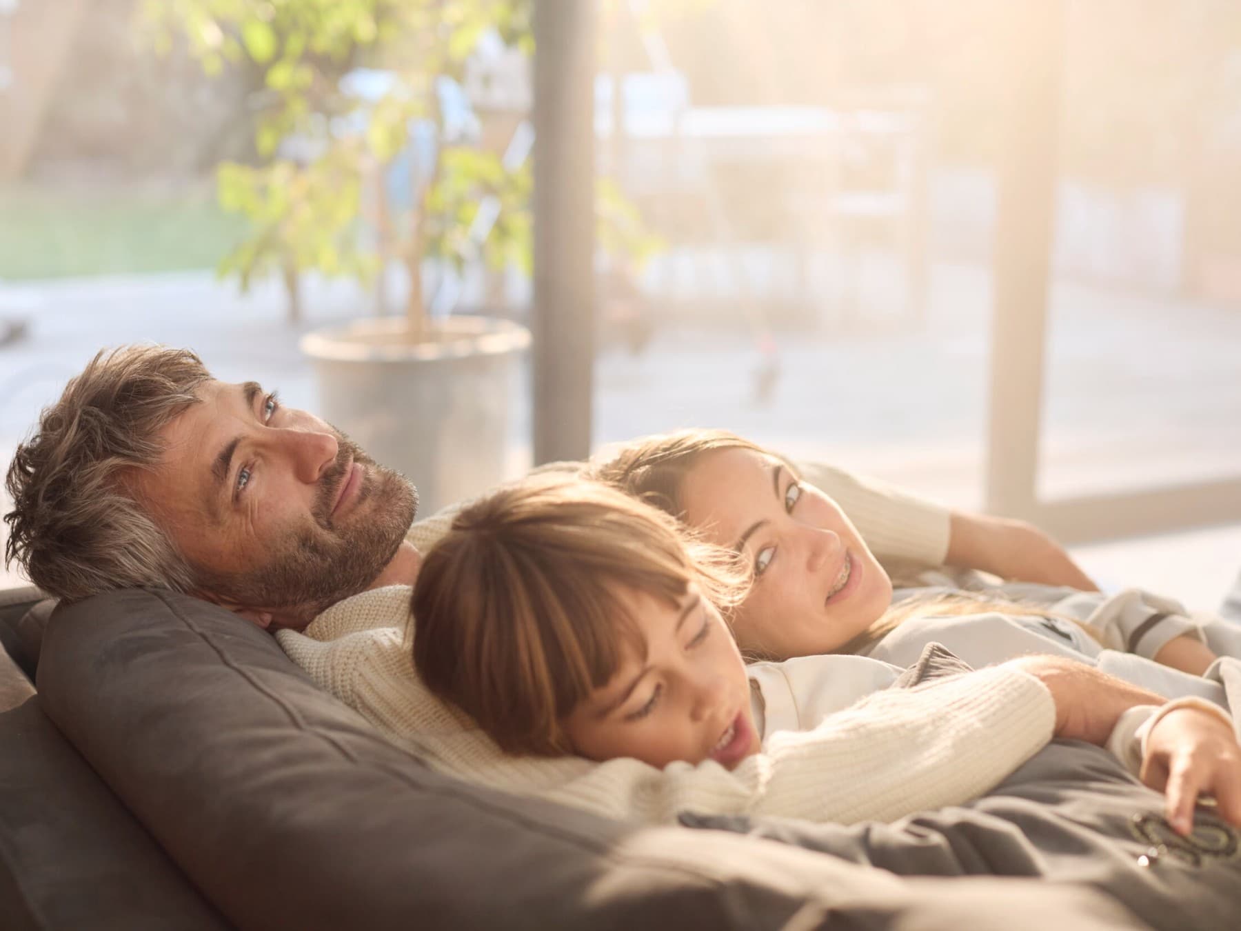Un père et ses deux enfants se détendent sur un canapé, la porte vitrée en arrière-plan offre une vue sur la terrasse.