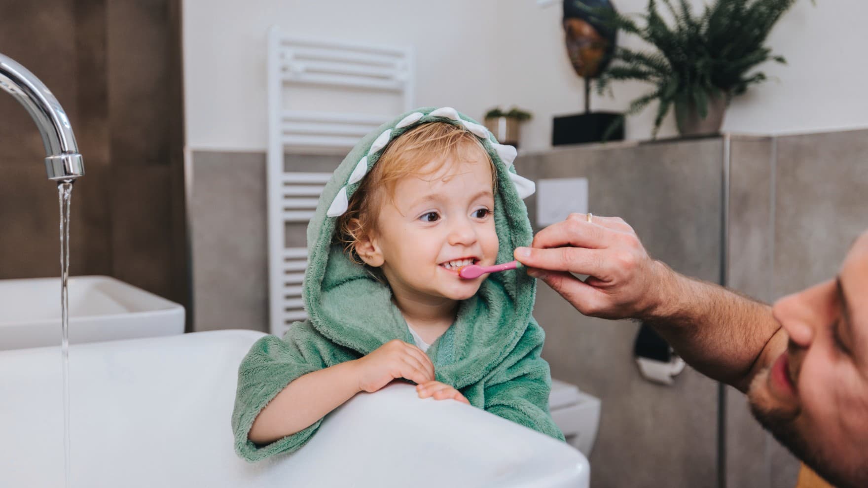 Un père brosse les dents de son enfant avec un sèche-serviettes chauffant en arrière-plan.