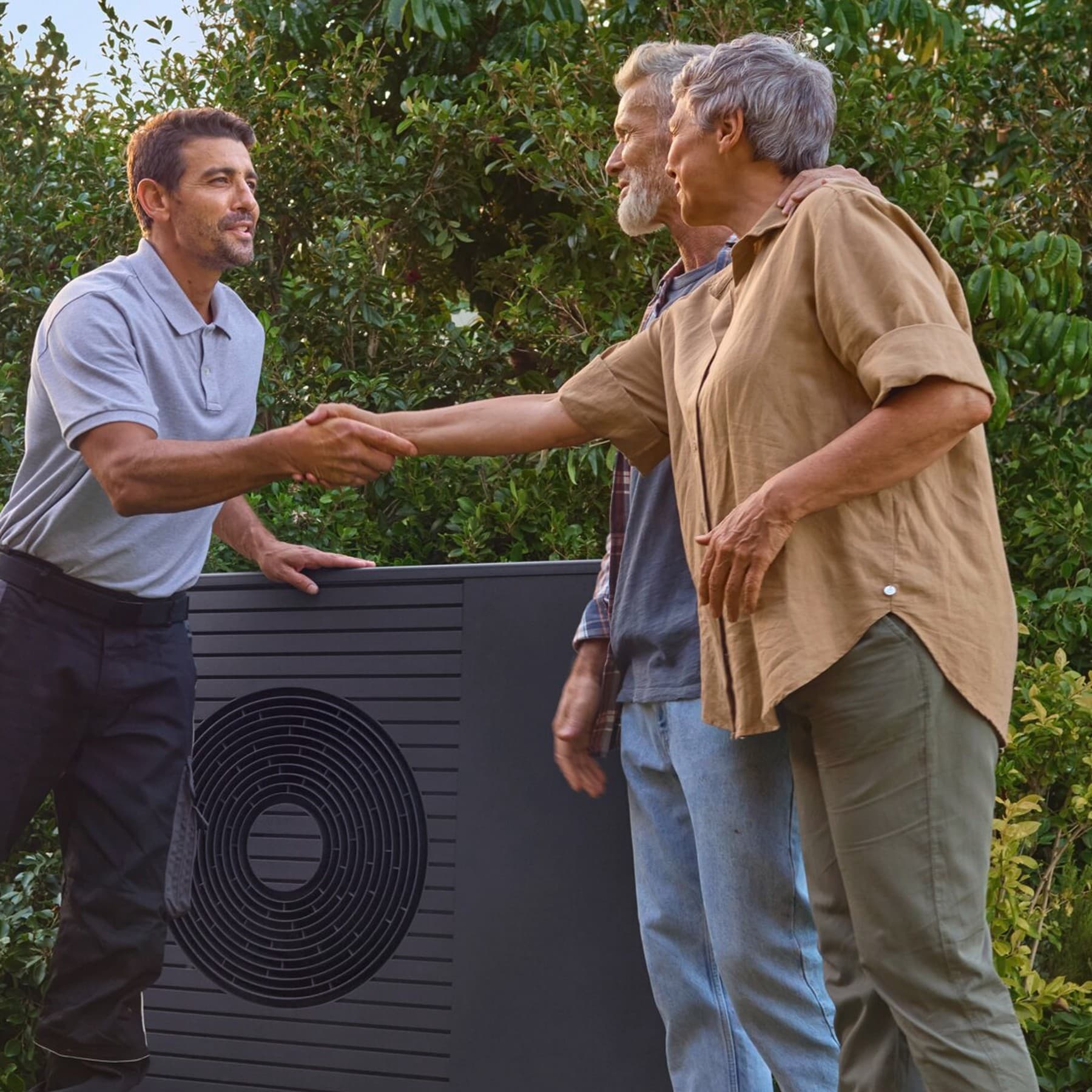 Een installateur en een echtpaar staan voor een Vaillant-warmtepomp in de tuin en schudden elkaar de hand.