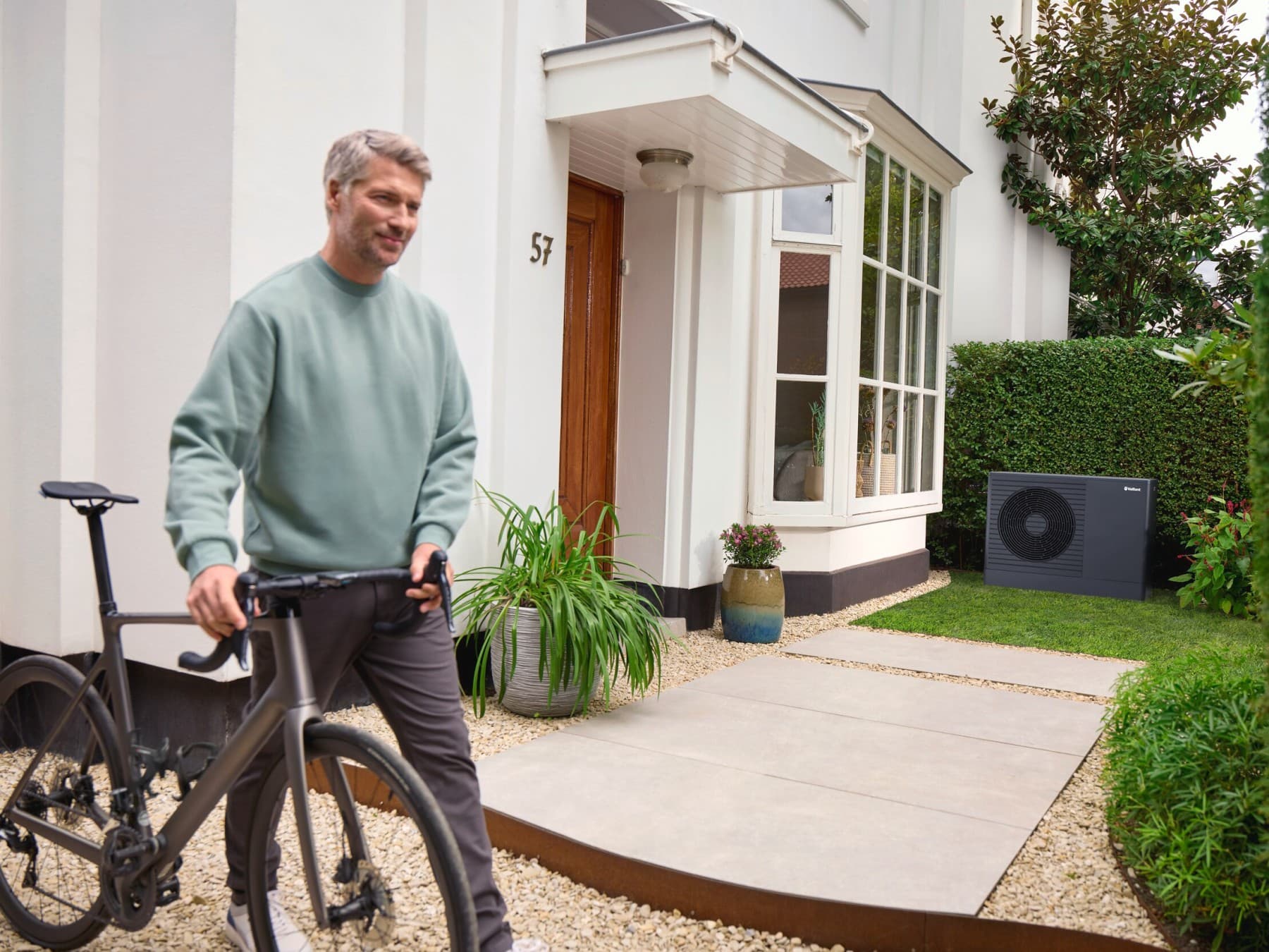 Un homme tient un vélo à la main ; à l'arrière-plan, on aperçoit une maison et une pompe à chaleur noire.