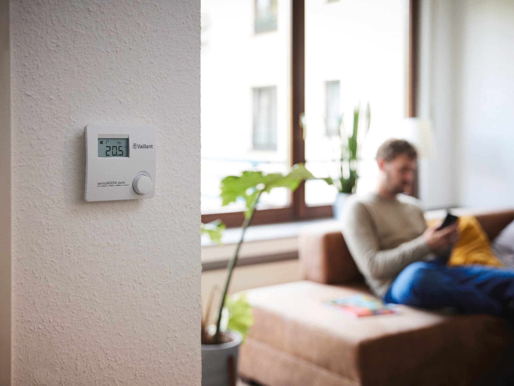 sensoROOM pure : régulation modulante fixée au mur, avec en arrière-plan un homme qui consulte son téléphone